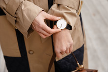 Street Style Fashion Details. Close Up, Young Fashion Blogger Wearing Autumn Trench Coat And A White And Golden Black Analog Wrist Watch. Checking The Time, Holding A Beautiful Brown Leather Purse.