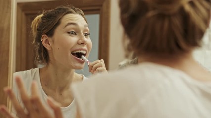 Sticker - An attractive smiling woman looking to the mirror while cleaning her teeth in the bathroom with brush and toothpaste