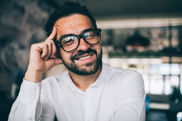 Portrait of cheerful male financial expert in casual shirt smiling at camera while waiting business partner for meeting in coworking space, happy Caucasian man in optical spectacles indoors