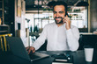© BullRun - Portrait of successful man in formal wear enjoying smartphone conversation with friend sitting at desktop with modern netbook and smiling at camera, male expert satisfied with distance job indoors