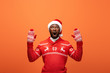 © LIGHTFIELD STUDIOS - shocked african american man in santa hat, mittens and Christmas sweater on orange background