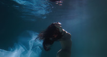Beautiful Girl Swims Underwater With Long Hair. Blue Or Gold Background Like Gold. The Atmosphere Of A Fairy Tale Or Magic. Diving Under The Water With A Shiny Cloth