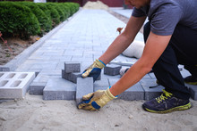 The Master In Yellow Gloves Lays Paving Stones In Layers. Garden Brick Pathway Paving By Professional Paver Worker. Laying Gray Concrete Paving Slabs In House Courtyard On Sand Foundation Base.