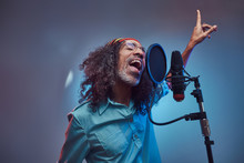 African Rastafarian Singer Male Wearing A Blue Shirt And Beanie Emotionally Writing Song In The Recording Studio. Isolated On A Blue Background.