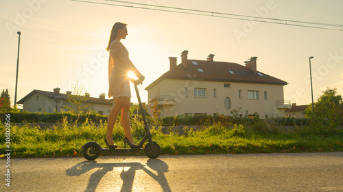 Fotomural LENS FLARE: Modern millennial woman commutes through suburbs on an e-scooter