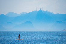 Woman Stand Up Paddle On Camboinhas Beach In Niteroi, Surfing With Christ The Redeemer Statue And The Mountains Of Rio De Janeiro In The Background, Clouds Separating The Mountains From The Blue Sea.