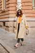 © mlasaimages - authentic street style portrait of an attractive woman wearing trench coat, sunglasses and slingback shoes, crossing the street.fashion outfit details perfect for autumn fall winter