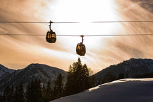 Ski Lift, Two Gondolas Against The Backdrop Of An Orange, Cloudy Sky. In The Background Visible Mountains Covered With Snow. Cable Car At Schladming Dachstein, Austria.
