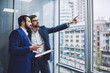© BullRun - Two caucasian business partners looking in window for building and thinking about making investment, successful entrepreneur pointing outdoors to male colleague standing near with paper folder in hand