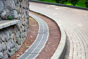 Wall Mural - grille of the drainage system on the curve pedestrian sidewalk of turn on the side of the sidewalk a storm channel with an iron grate and rock stone wall.