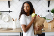 African girl standing on the kitchen holds a paper bag with food and eats an apple