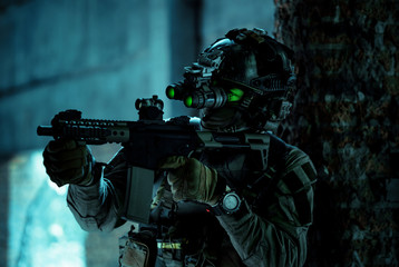 Man in uniform with machine gun and turned on night vision device inside broken building. Airsoft soldier with green light on face in night building