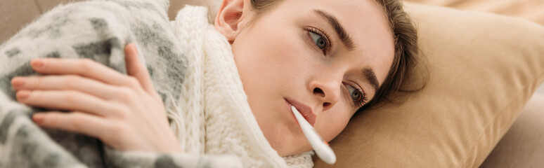 panoramic shot of sick woman lying on sofa under blanket and measuring temperature