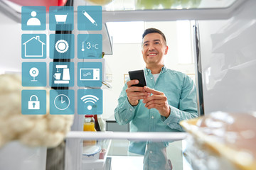 healthy eating, technology and diet concept - happy middle-aged man with smartphone making list of necessary food from fridge at kitchen over blue and white smart home app icons