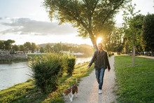 Young Man Takes His Beloved Dog For A Walk In The Park At Sunset - Millennial In A Moment Of Relaxation With His Four-legged Friend