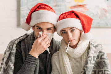 diseased couple wearing santa hats and covered with blanket looking at camera