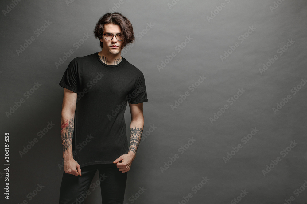 Hipster handsome male model with glasses wearing black blank t-shirt and black jeans with space for your logo or design in casual urban style - obrazy, fototapety, plakaty 