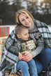 © sofiko14 - Mom and son enjoying snowy winter day outdoors, sitting on the sledge, hugging, covered with plaid and holding Christmas gifts