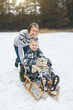 © sofiko14 - Beautiful family of young mother and son enjoying snowy winter day outdoors having fun sledging. Boy is sitting on the wooden decorated sledge with presents, while mom is pushing him