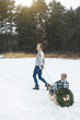 © sofiko14 - Winter family fun holidays. Happy mom in knitted sweater and jeans carries her little son on a wooden sled decorated with Christmas wreath in a winter snowy forest. Merry christmas and happy new year