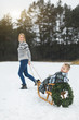 © sofiko14 - Winter family fun holidays. Happy mom in knitted sweater and jeans carries her little son on a wooden sled decorated with Christmas wreath in a winter snowy forest. Merry christmas and happy new year