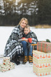 © sofiko14 - Little boy and mother sliding in the winter forest. Mom and son sitting on the sledge, covering by the checkered plaid and drinking hot drinks. Decorated present gifts around them
