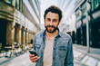 © BullRun - Portrait of handsome young man with beard smiling at camera while dialing number on phone standing outdoors on street near shopping center in city.Good looking caucasian customer holding mobile