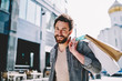 © BullRun - Portrait of happy hipster guy enjoying big sales during shopping in black friday.Positive bearded young man customer with paper bags smiling at camera standing outdoors on street near mall
