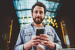 © BullRun - Half length portrait of handsome caucasian bearded guy holding mobile phone connected to 4G for blogging, casually dressed male hipster using modern smartphone outdoors satisfied with updates.