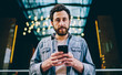 © BullRun - Half length portrait of handsome bearded man holding smartphone connected to 4G checking notification, confident millennial man blogger using mobile phone for share and chatting in social networks.