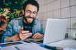 © BullRun - Positive caucasian hipster guy in eyewear laughing at content from social networks using mobile phone for browsing, cheerful male freelancer having fun during remote job  chatting on cellular.