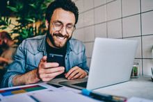 Positive Caucasian Hipster Guy In Eyewear Laughing At Content From Social Networks Using Mobile Phone For Browsing, Cheerful Male Freelancer Having Fun During Remote Job  Chatting On Cellular.