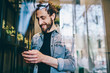 © BullRun - Cheerful bearded hipster guy chatting with friends in social networks holding smartphone in hands.Positive young man blogger sharing funny publication on own website on cellular standing in coworking