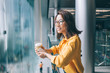 Young Asian woman with coffee and cellphone leaning on railing smiling and daydreaming