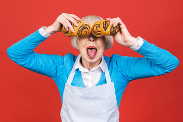 Portrait of pretty charming cheerful senior aged woman with wrinkle showing gesturing sweet homemade bakery tasty bun isolated on red background, specialty of the house.