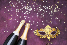 Two Champagne Bottles, Golden Carnival Mask And Confetti Stars On Purple Background. Flat Lay Of Christmas, New Year, Purim, Carnival Celebration Concept. Copy Space, Top View.