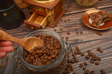 The Girl's Hand Takes A Wooden Spoon Of Coffee Beans From A Glass Jar, A Coffee Grinder, Pieces Of Chocolate On A Wooden Background. Close Up.