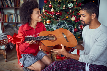 Wall Mural - Couple in front of decoration Christmas tree. man surprise woman a with a guitar gift for Christmas.