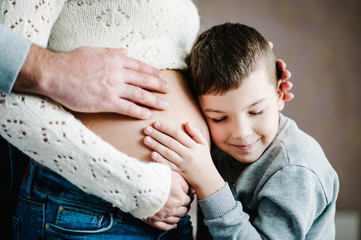 Son is listening to his beautiful pregnant mother tummy and smiling. Woman and loving man, boy hugging tummy at home. Parenthood concept. Baby Shower. Second pregnancy. Brother.