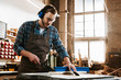 © LIGHTFIELD STUDIOS - handsome woodworker in protective headphones and apron holding plank near circular saw