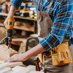 cropped view of carpenter in apron holding hammer drill near wooden plank