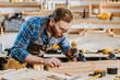 © LIGHTFIELD STUDIOS - selective focus of carpenter in safety glasses and apron holding hammer drill and blowing on sawdust near wooden plank