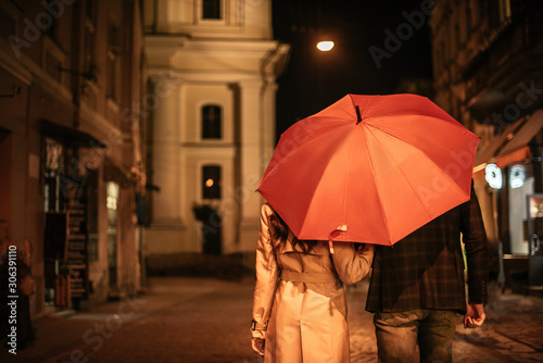 Fotografía back view of couple in autumn outfit walking under umbrella along evening street