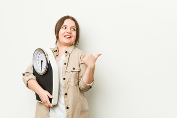 Sticker - Young curvy woman holding a scale points with thumb finger away, laughing and carefree.