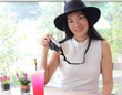 © Phuttharak - Beautiful Asian tourist woman wearing black hat  sitting in coffee shop with red drink on table holding her camera