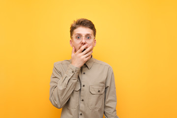 Portrait of shocked young man in shirt on yellow background, covered open mouth with hand surprised and looking into camera. Surprised nerd isolated on yellow background.