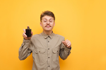 A funny guy in a shirt stands with a bottle of perfume in his hand against a yellow background and enjoys the smell with his eyes closed. Young male nerd uses perfume, isolated.