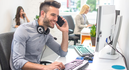 Wall Mural - Handsome businessman using a computer, talking on the mobile phone and smiling while working in office