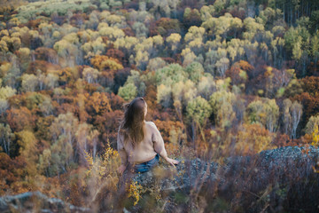 Young woman sitting on cliff's edge, looking at beautiful autumn forest.