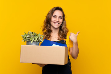 Young blonde girl making a move while picking up a box full of things with thumbs up because something good has happened